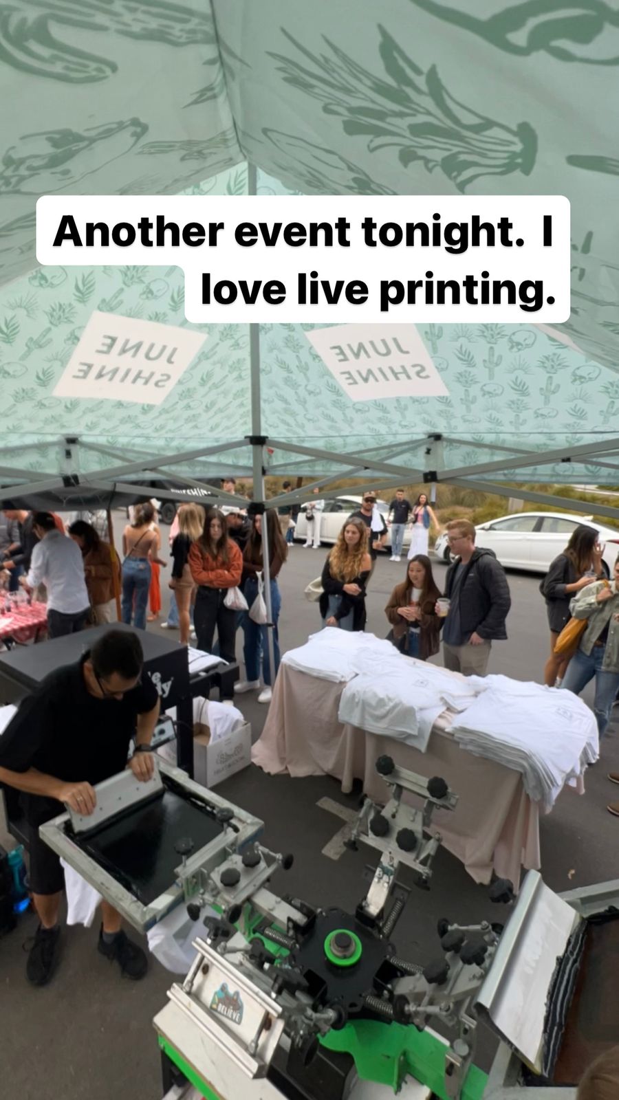 Guests gathered around a live printing tent at an outdoor welcome party with stacks of white garments