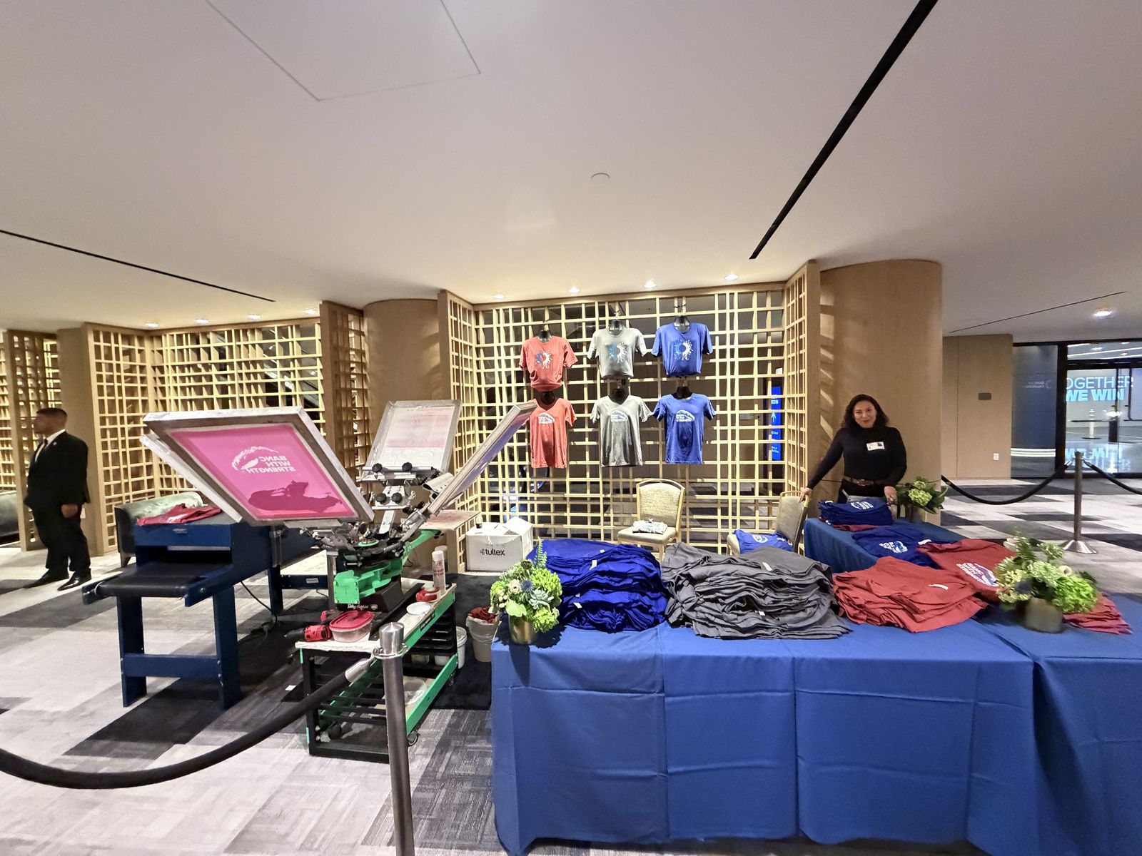 Merch station inside a hotel venue with shirts displayed on a wooden lattice wall and folded stacks on navy linens
